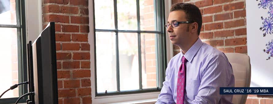 Online finance degree student studies on laptop by window. With the text Saul Cruz '16 19 MBA