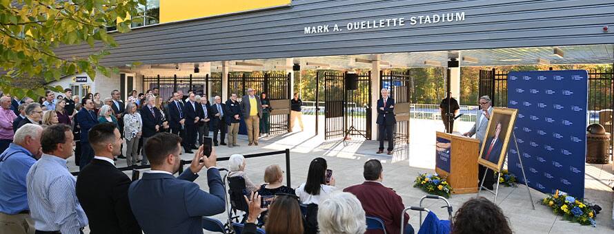 A group gathered for a surprise dedication ceremony in front of the newly renamed Mark A. Ouellette Stadium on the SNHU campus.
