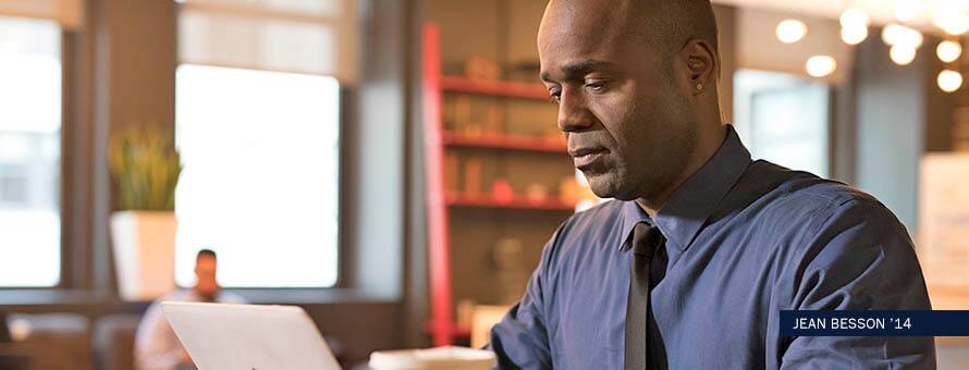 IT degree student wearing a blue shirt and black tie, studies at laptop in library. 