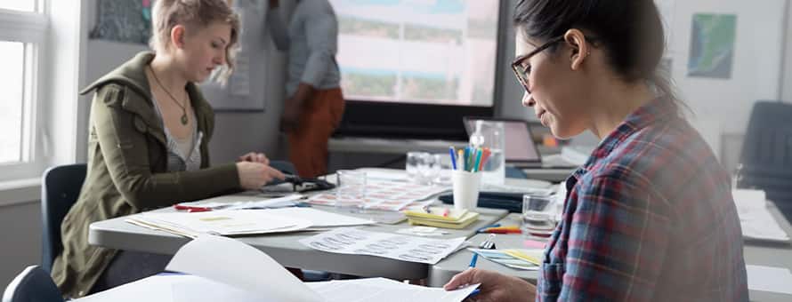 A public administration professional at a table, reviewing documents with a pen in hand, while two colleagues do their work.