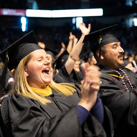 Shelly and Sal Villa wearing their cap and gowns, clapping and cheering at SNHU's commencement.
