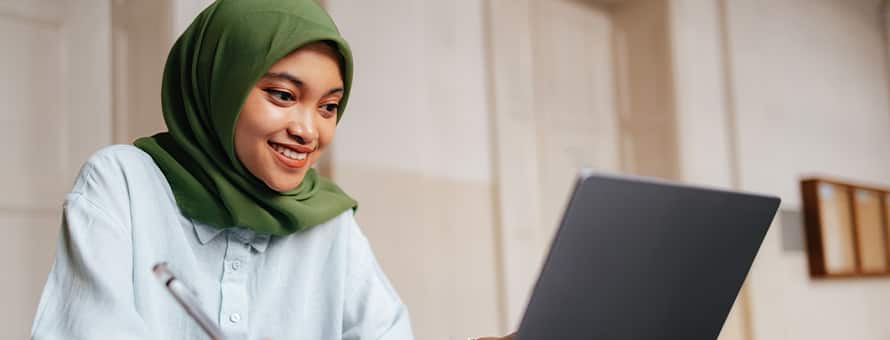 A smiling woman wearing a green hijab and a button-down shirt sitting at a laptop with a pen in hand.