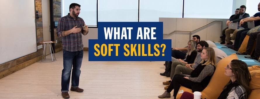 A man in a plaid shirt speaking to a group of people sitting on beanbags and tiered steps and text what are soft skills?