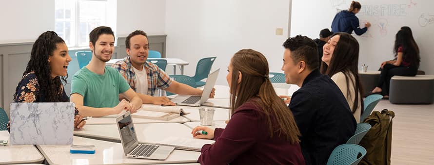 A group of six college students at a large table covered with notebooks and laptop computers.