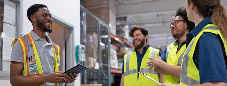 A supply chain manager holding a tablet, speaking with three other professionals wearing fluorescent yellow vests and working in a warehouse.