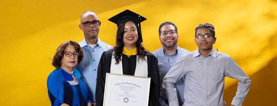 Brendaliz De Los Santos, center, dressed in a graduation cap and gown and holding her SNHU diploma, surrounded by 4 family members who supported her in college.
