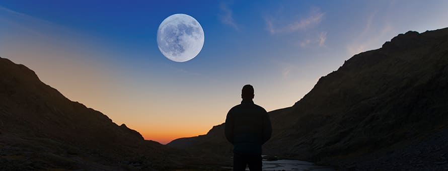 The silhouette of a man standing between two hills at dusk looking  at the moon.