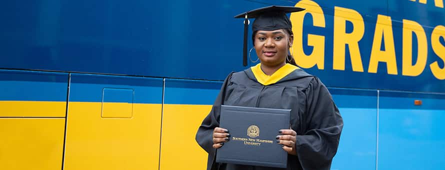 SNHU graduate from Florida in their cap and gown, holding their diploma in front of the SNHU bus
