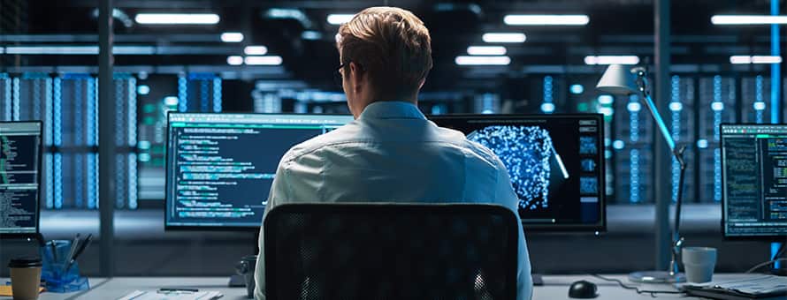 The back of a professional with a master's in cybersecurity at work, sitting at a desk in a large office building with two monitors in front of him.