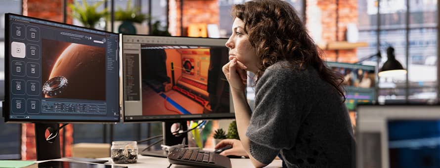 A game designer sitting in front of two monitors with a video game development software open, showing a planet and the inside of a spacecraft.