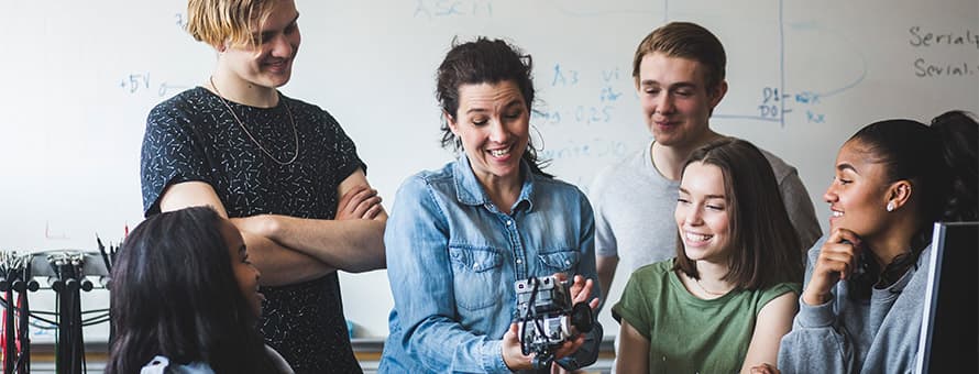 A STEM educator holding a piece of robotics as 5 students watch.