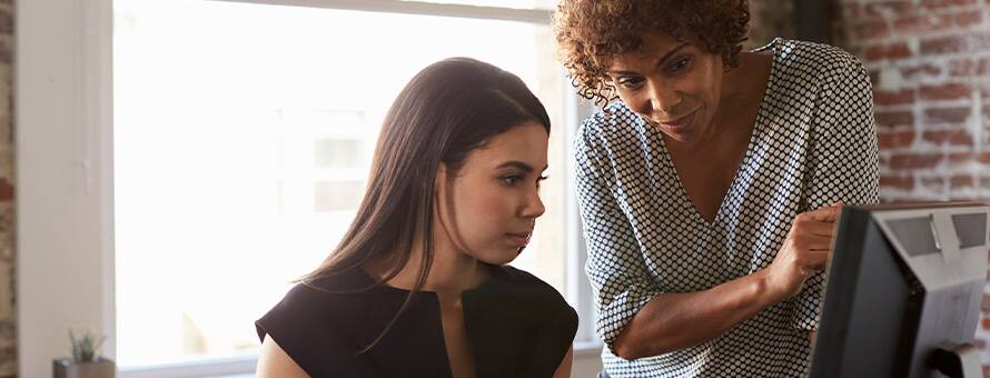 One woman sitting look at a desktop with another woman standing over her looking at the desktop