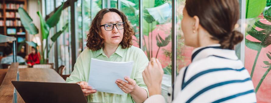 A career advisor holding a student’s resume, listening to the student talk about what she wants to include in a cover letter.