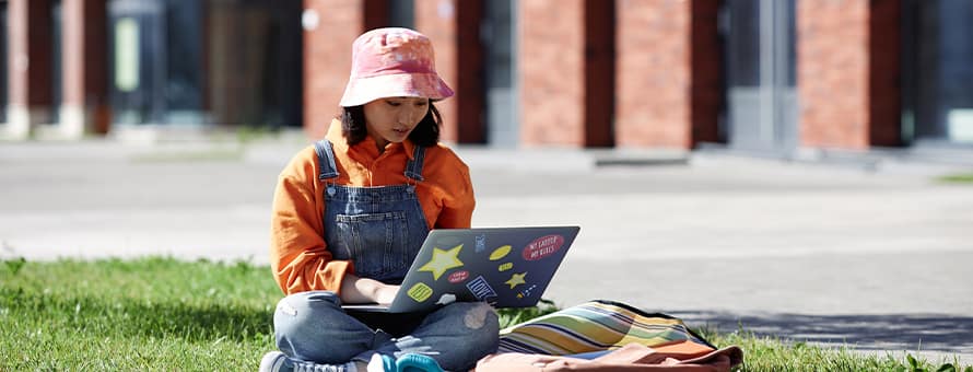 A Gen Z college student sitting in a grassy area while doing schoolwork on her laptop.
