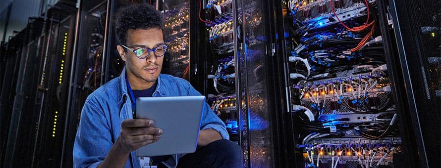 A student exploring what cyber security is, holding a tablet and standing in front of large machines at his internship.