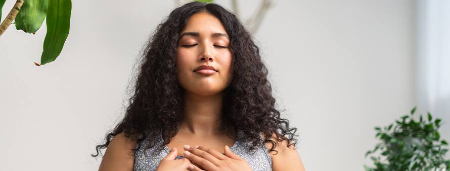 A woman surrounded by plants, meditating with her eyes closed and hands over her chest