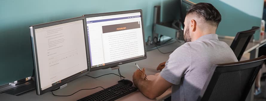 A transfer student sits in a library working on online coursework using two computer monitors.