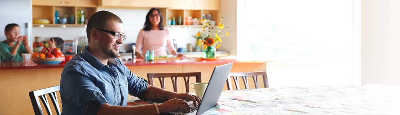 A student working on his laptop in a kitchen with his family nearby