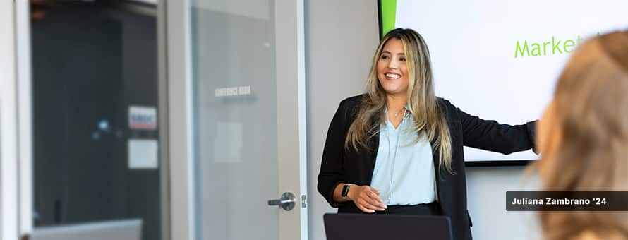 Juliana Zambrano '24, dressed professionally in a blouse and blazer, presenting in front of a screen in a board room.