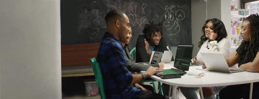 A group of students in a study room working on laptops together and smiling.