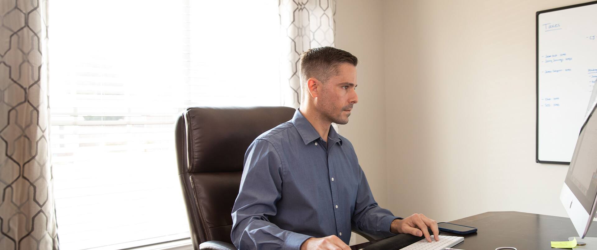 SNHU graduate Blake Venable sitting at a desk with a computer monitor and keyboard with a window and whiteboard on the walls behind him.