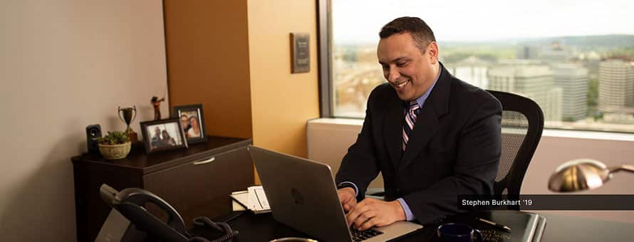 Stephen Burhart, wearing a dark suit, typing on a laptop in an office with a large window behind him overlooking city buildings.