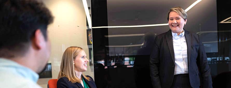A professionally dressed woman smiles at her coworkers in an office setting.