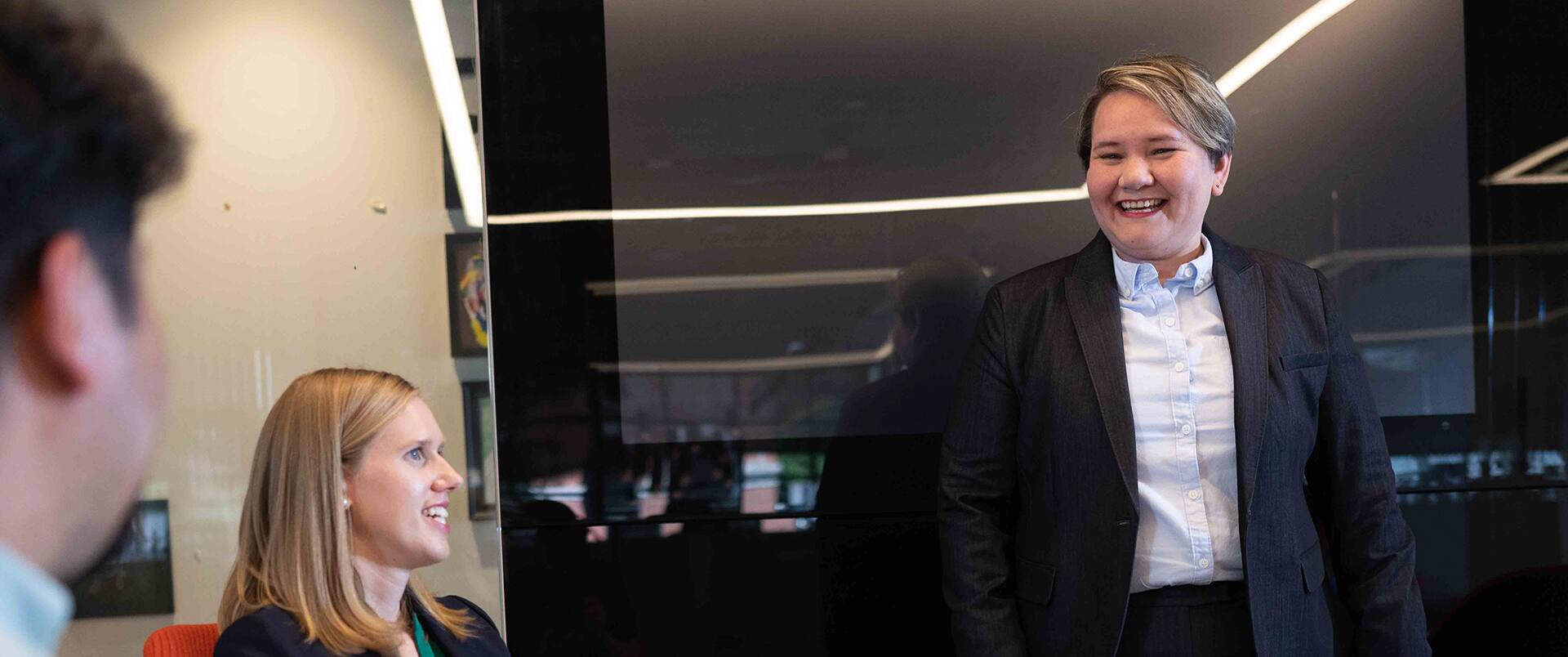 A woman with short hair wearing a dark blazer and speaking to a man and woman in a conference room.