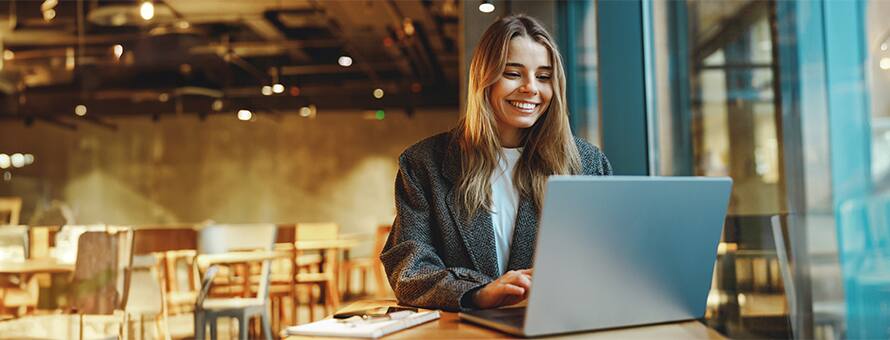 A woman sitting in a cafe, using a laptop to research entry-level jobs with a notebook open beside her.