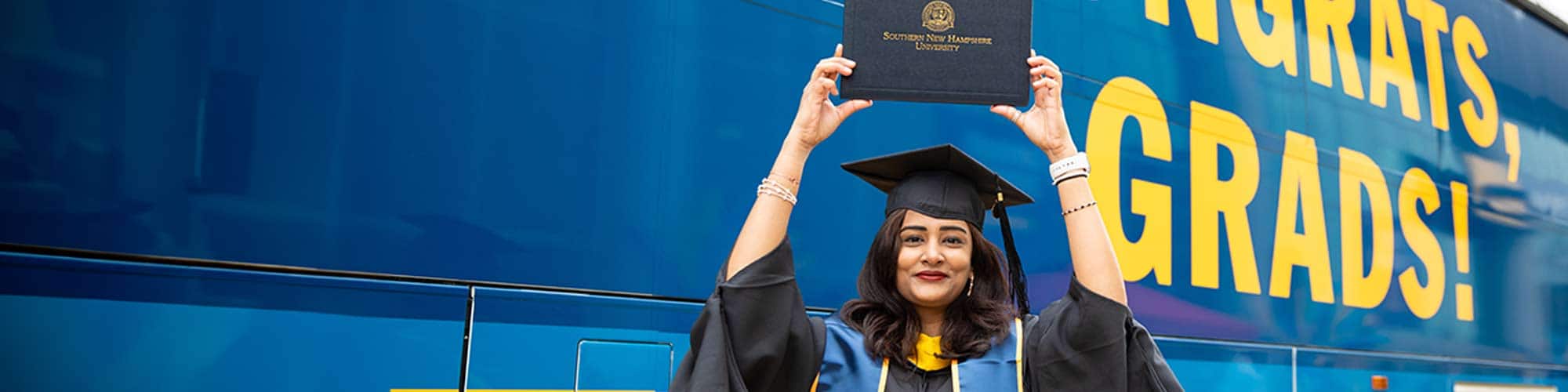 An SNHU grad in a cap and gown holding her diploma up in front of an SNHU branded bus