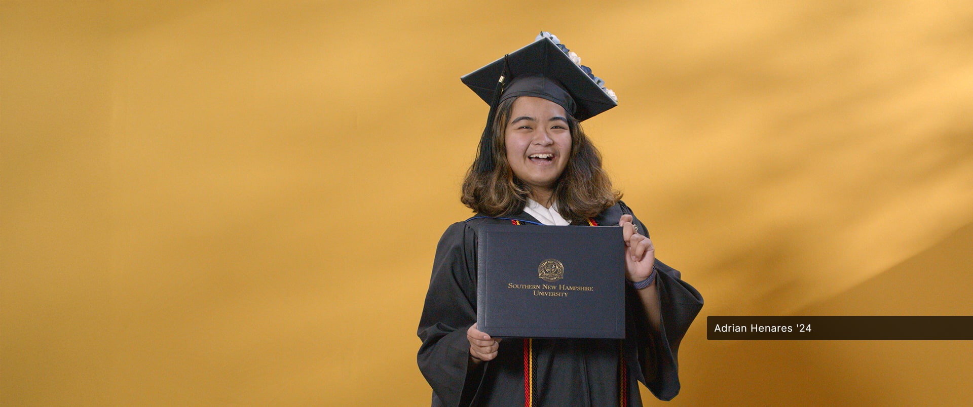 Adrian Henares, who earned her online bachelor's in communication from SNHU in 2024, wearing her cap and gown and holding her diploma in front of a yellow backdrop.