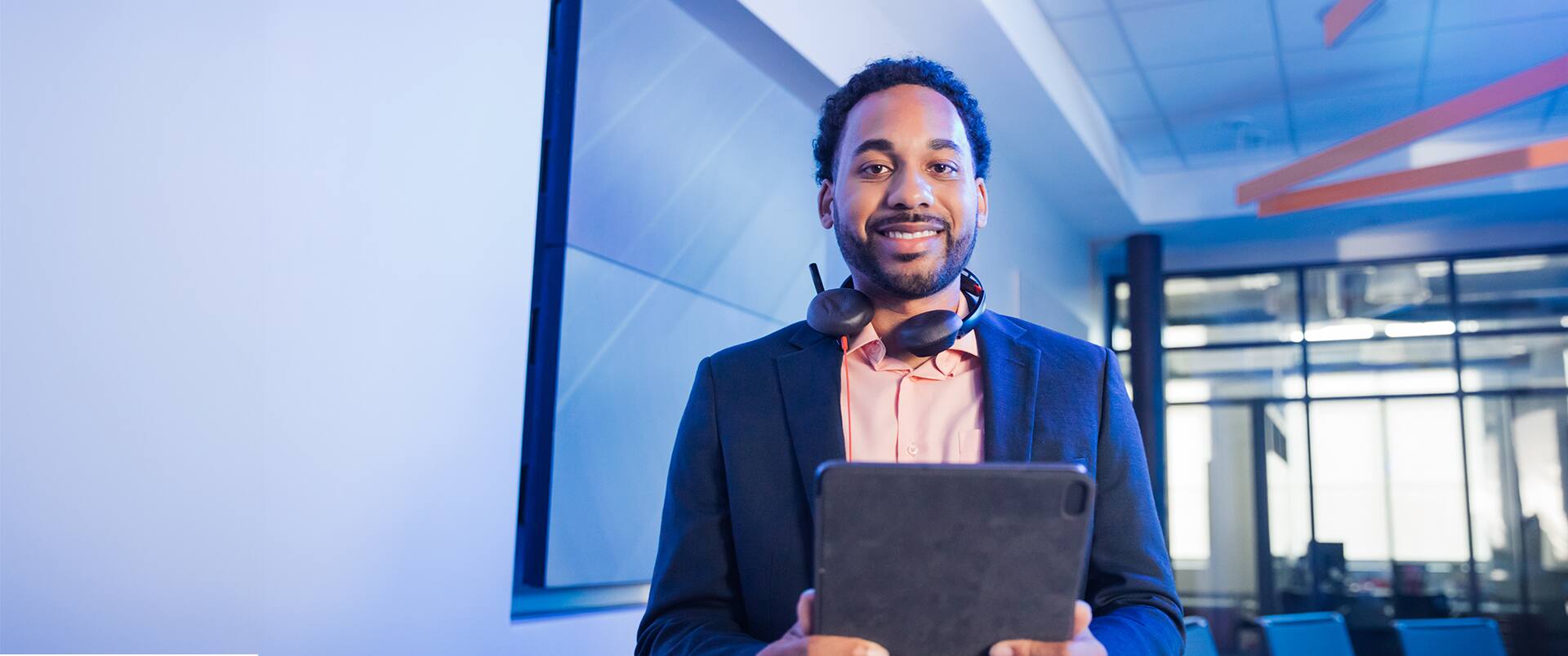A man standing next to a glass wall with headphones around his neck and holding a tablet computer.