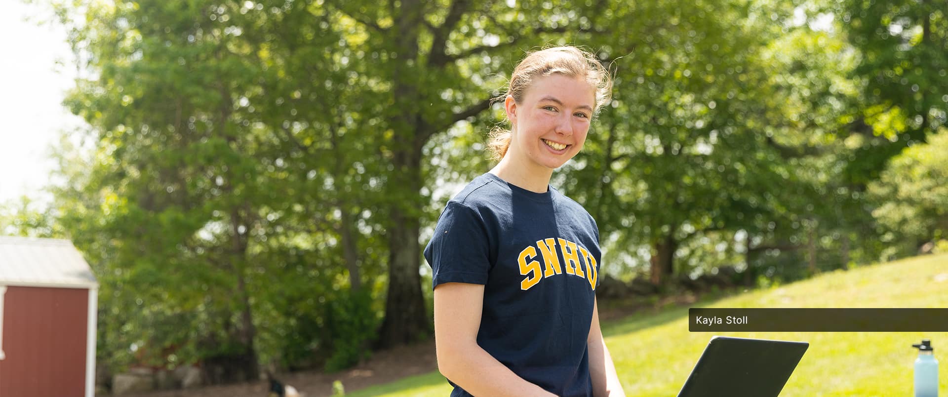 Kayla Stoll, who is earning her online degree in environmental science at SNHU, wearing a blue and yellow SNHU T-shirt and typing on a laptop with a large green tree in the background.