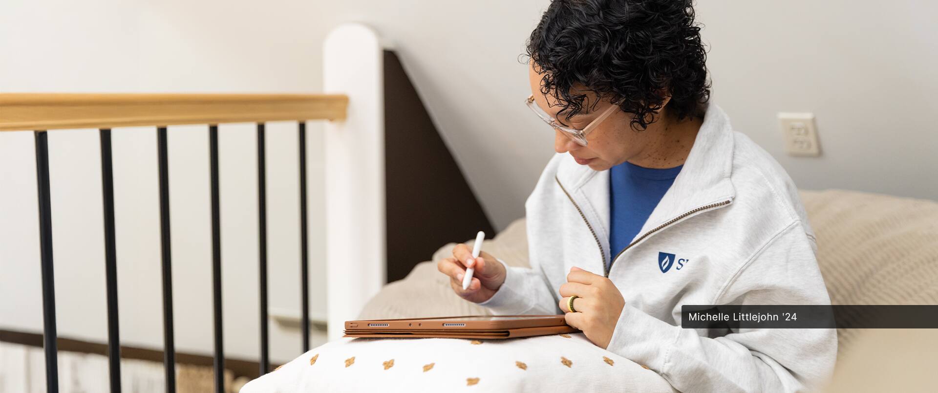 Michelle Littlejohn, who earned her online bachelor's in graphic design from SNHU in 2024, wearing a white SNHU sweatshirt and using a smart pen to draw on a tablet computer resting on pillow.