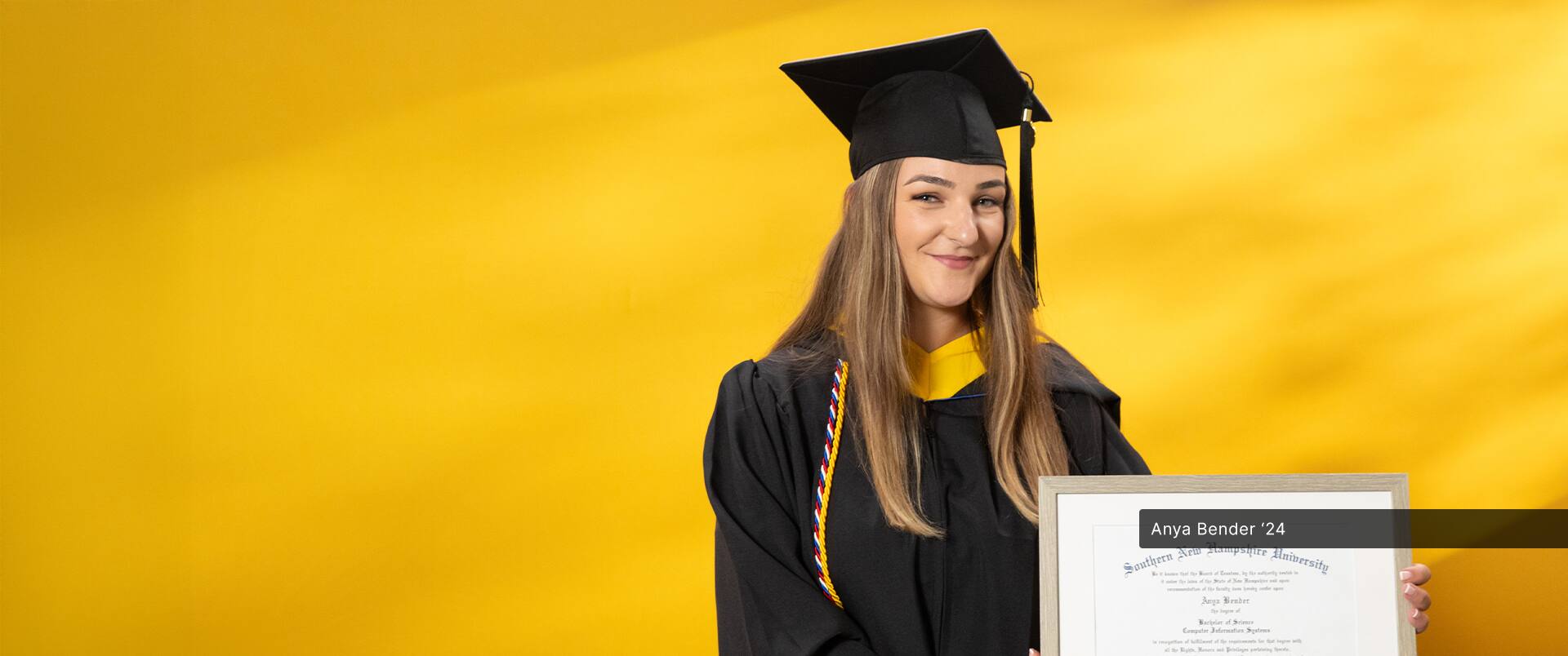 Anya Bender, who earned her online bachelor's in computer science from SNHU in 2024, wearing her cap and gown and holding her diploma in front of a yellow backdrop.