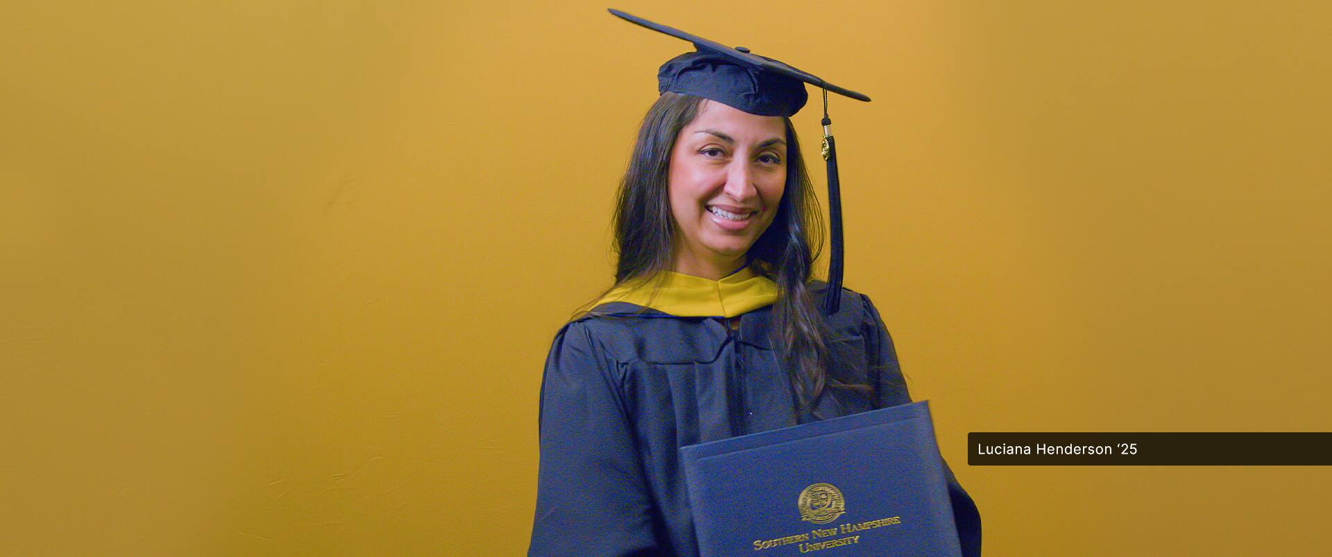 SNHU graduate, Luciana Henderson '25G, wearing cap and gown while holding her diploma.