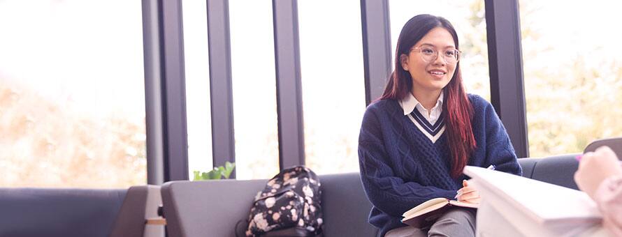 A student sitting at a couch in a study hall while talking to another individual across from her.