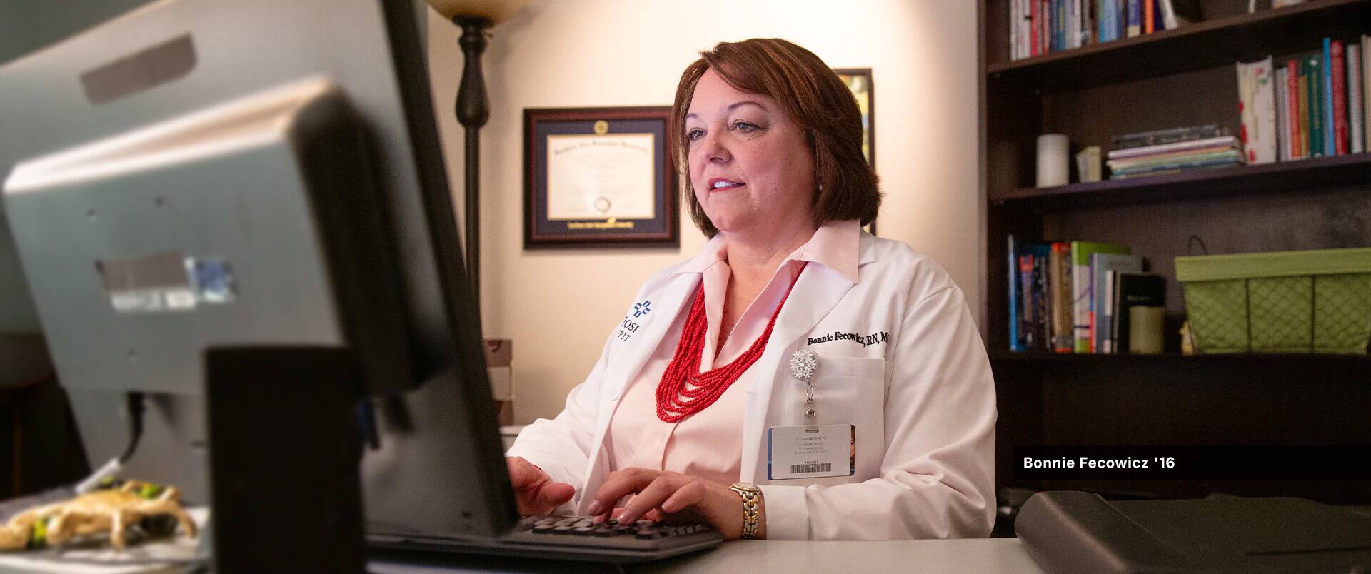 Bonnie Fecowicz, who earn her degree from SNHU, wearing a white lab coat, working on a desktop  computer at a desk with her framed SNHU diploma on the wall behind her.