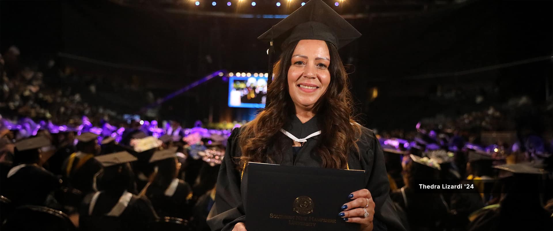 Thedra Lizardi, who earned her associate in liberal arts from SNHU in 2024, wearing her cap and gown and holding her diploma with the large commencement crowd seated in the background.