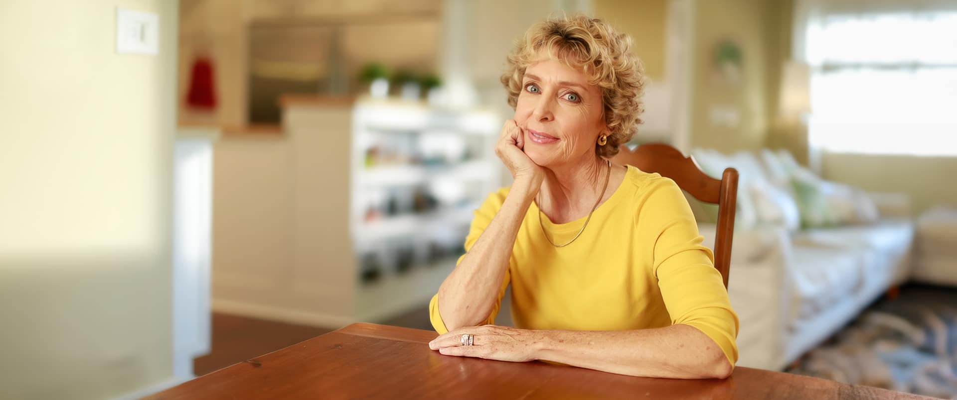"Susan Ryan, who earned her degree from SNHU, wearing a bright yellow shirt sitting at a  dining room table and resting her chin in her right hand."