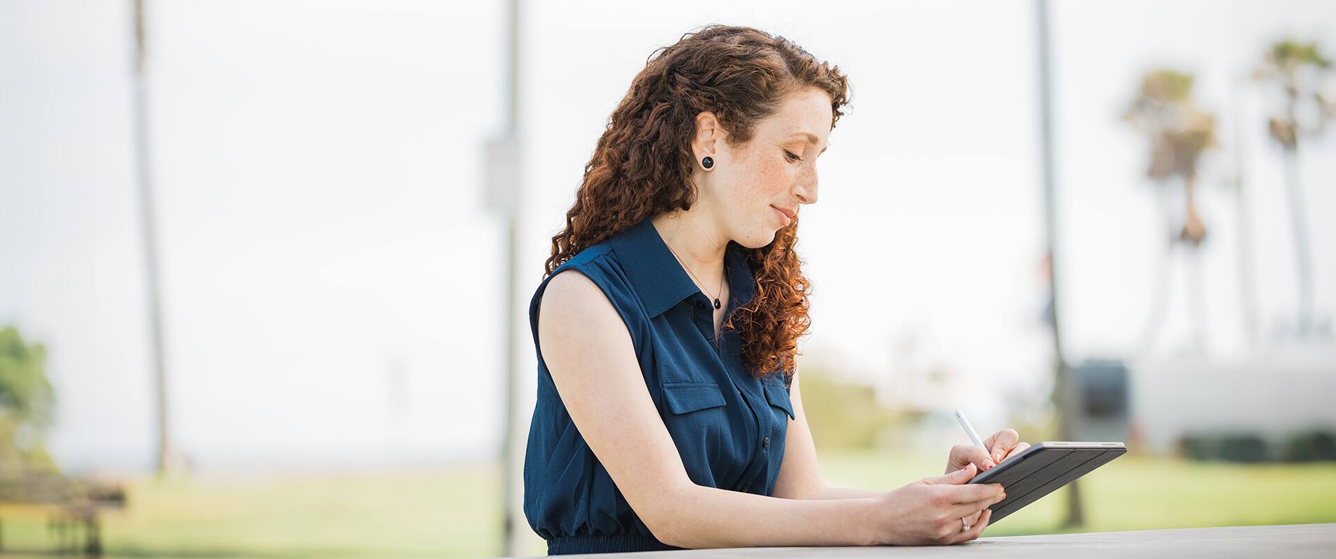 Mariel Embry, who earned her degree from SNHU, sitting at an outdoor table using a stylus and writing  on a tablet computer.