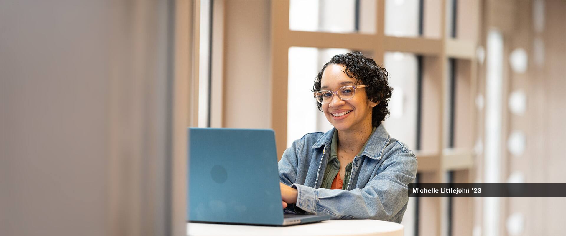 Michelle Littlejohn, who earned her online bachelor's in graphic design and media arts from SNHU in 2023, wearing a denim jacket sitting at a small table with her open laptop.