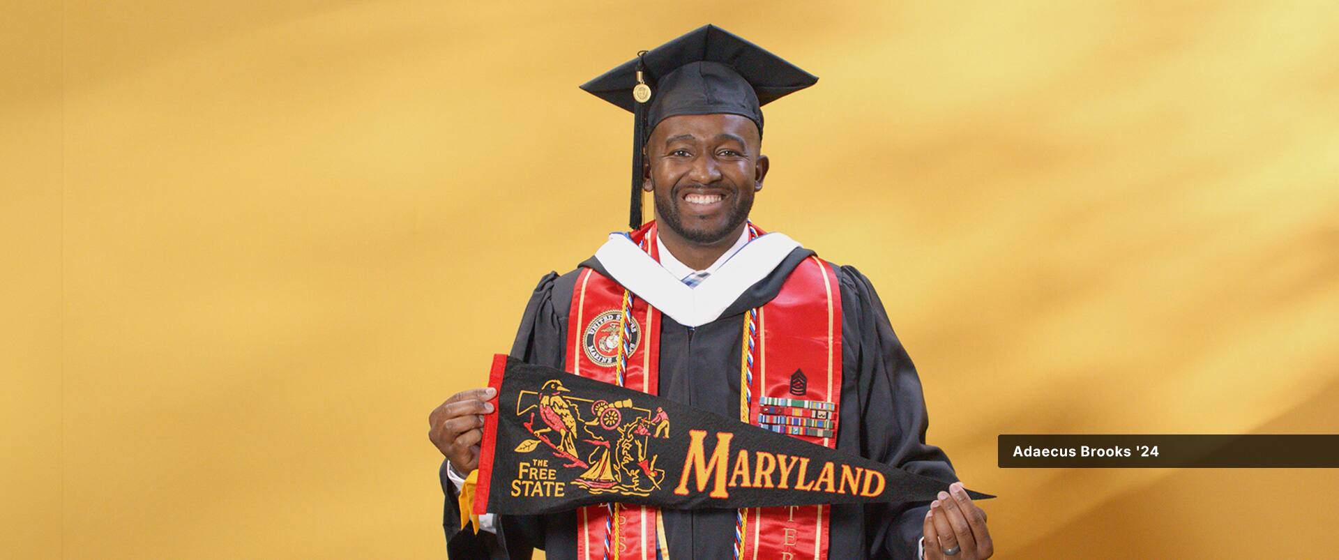 Adaecus Brooks, who earned his online bachelor's in graphic design and media arts from SNHU in 2024, wearing his cap and gown and holding a Maryland pennant.