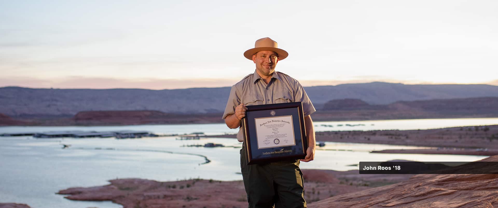 John Roos, who earned his history degree in 2018, wearing his park ranger uniform and hat and holding his framed SNHU degree with a lake and small mountains in the distance behind him.