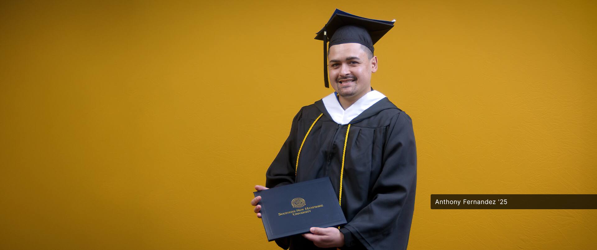 Anthony Fernandez, who earned his bachelor's in political science from SNHU in 2025, wearing his cap and gown and holding his diploma in front of a yellow backdrop.