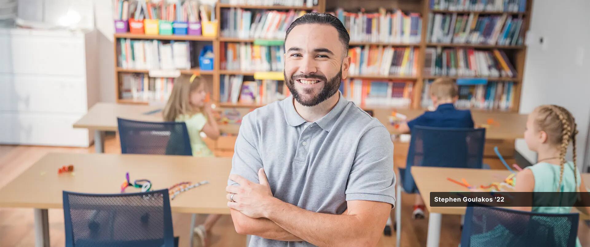 Stephen Goulakos, a 2022 psychology degree online graduate, standing in the middle of a classroom with young children playing at their desks.
