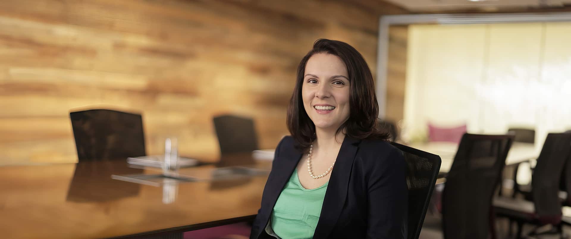 Jessye Giley wearing a light green blouse, dark blazer and pearl necklace sitting in a wood paneled conference room with a large table and several chairs in the background.