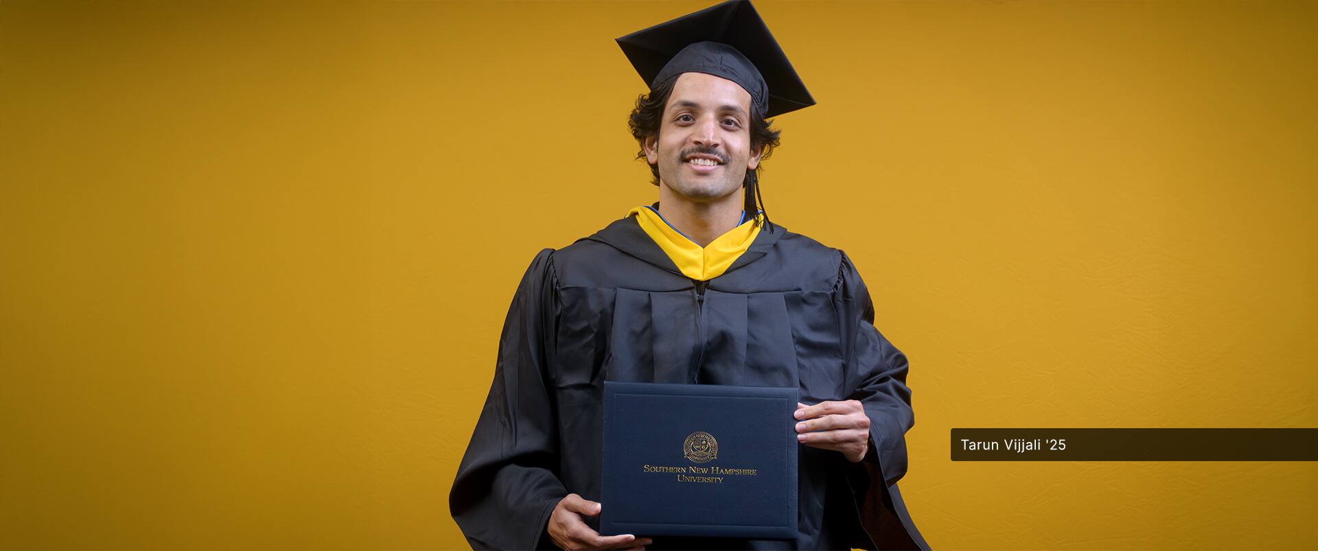 Tarun Vijjai, who earned his bachelor's in business administration with a concentration in management information systems, wearing his cap and gown and holding his diploma in front of a yellow backdrop.