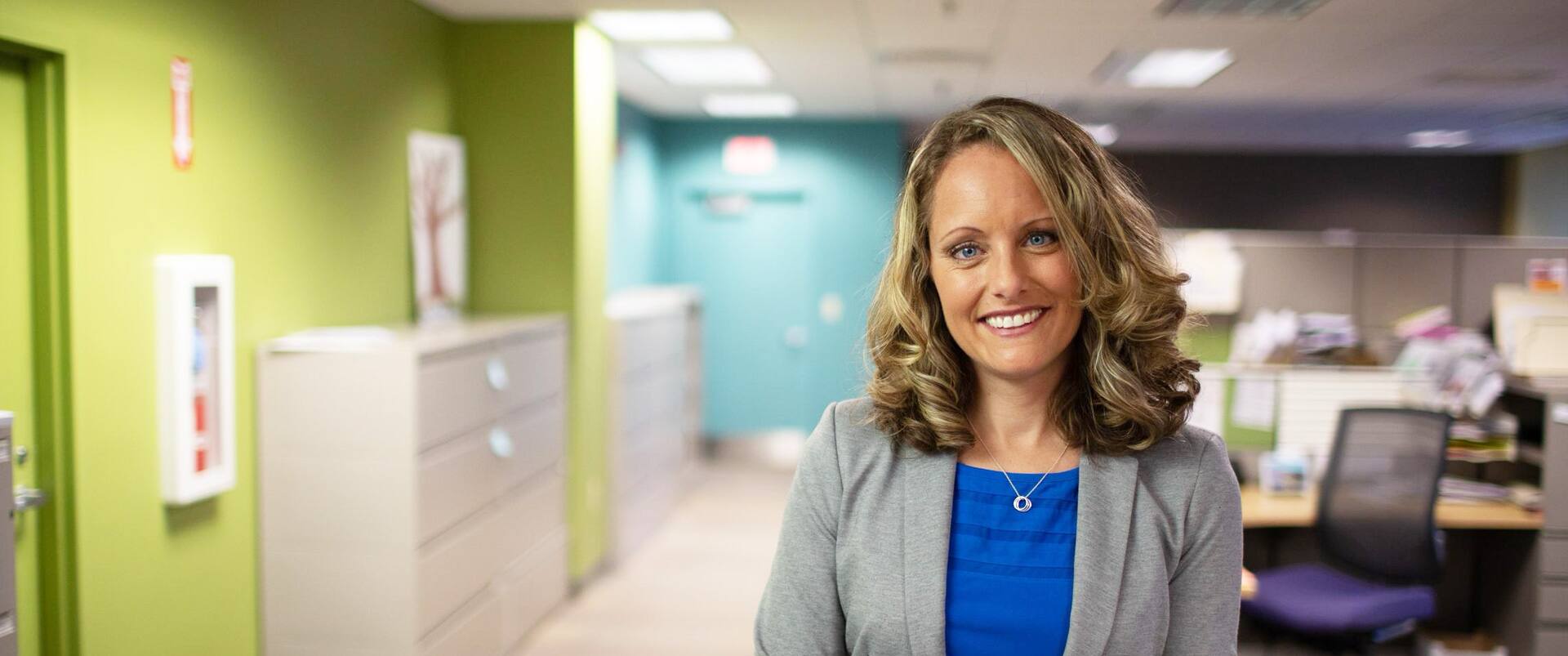 Kerry Smith, who earned her degree from SNHU in 2012, wearing a blue shirt and grey blazer standing in an office with a large filing cabinet and office cubicle in the background