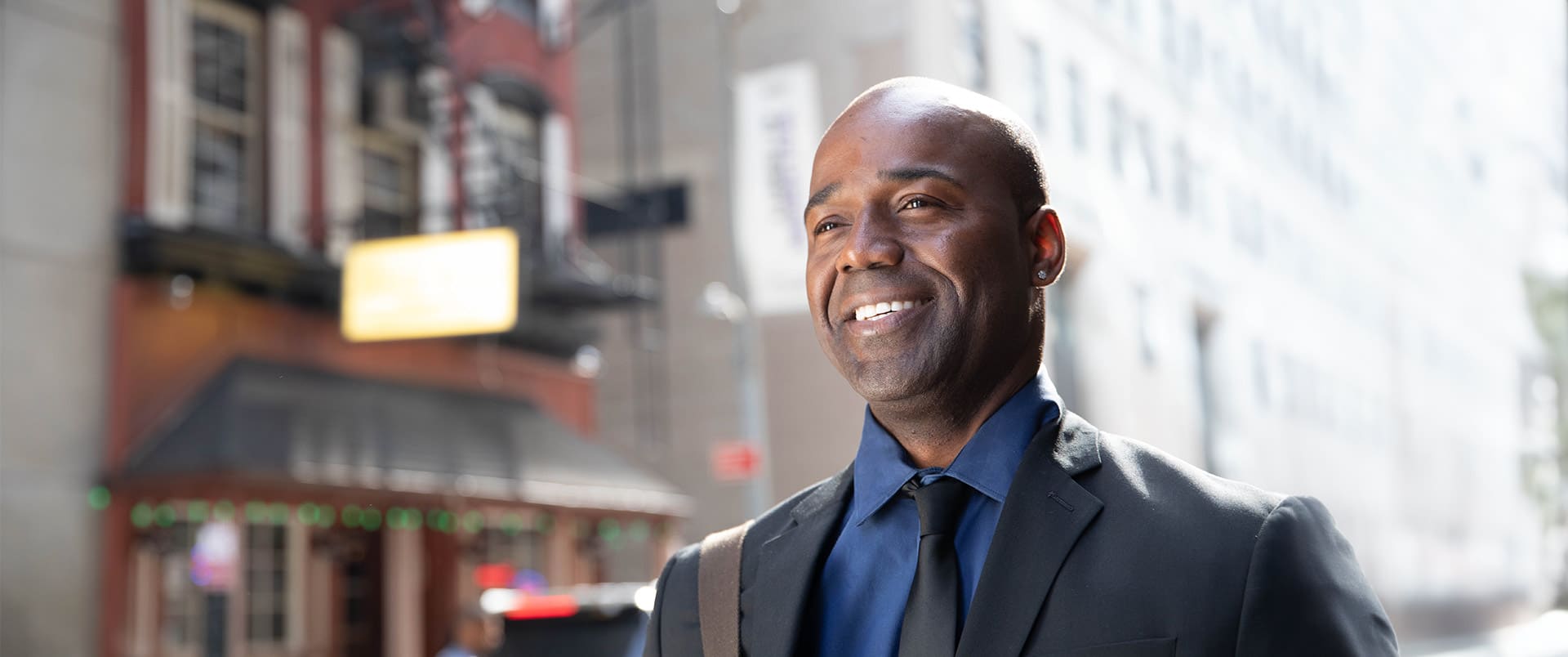 Jean Besson, who earned his degree from SNHU in 2014, wearing a suit and tie walking down a city street with a shoulder bag on his right shoulder.